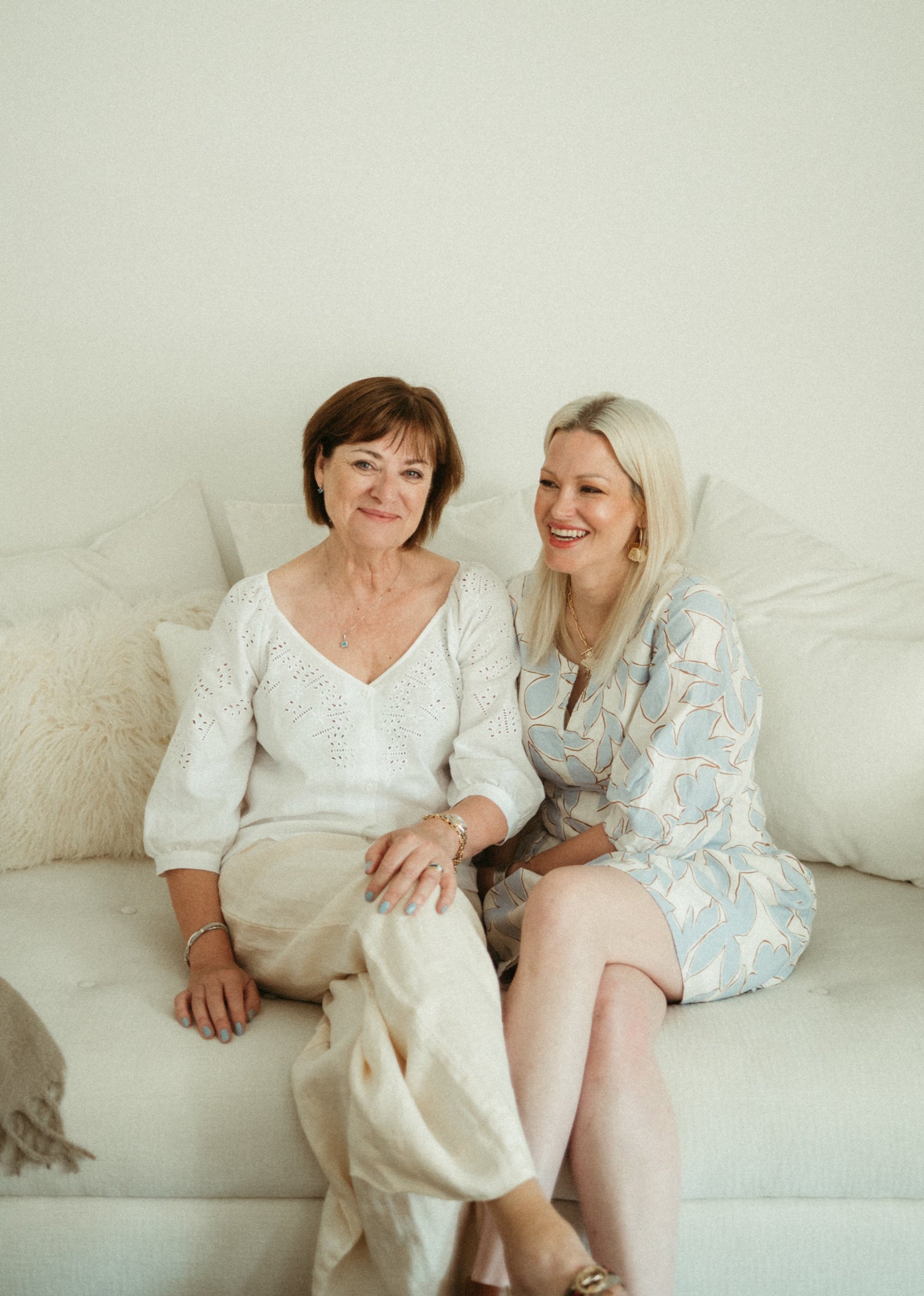 Two women sitting on a white couch in a softly lit room.