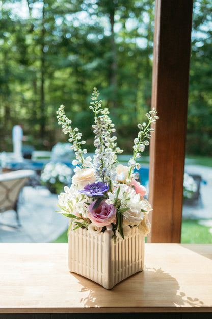 Floral arrangement in a decorative pot on a table with a blurred outdoor background