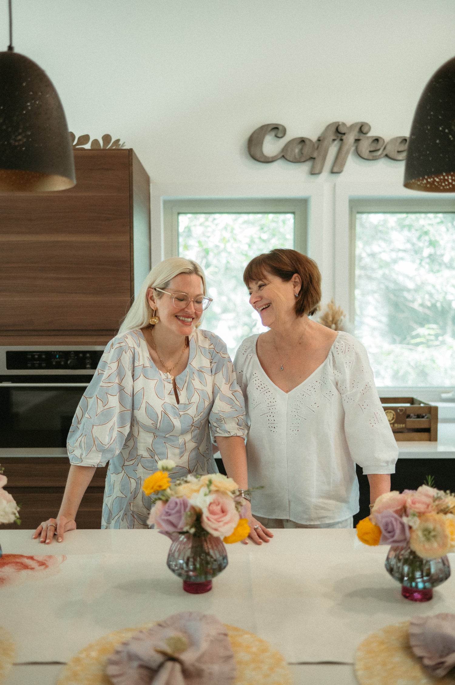 Two women standing in a kitchen with floral arrangements on the counter.