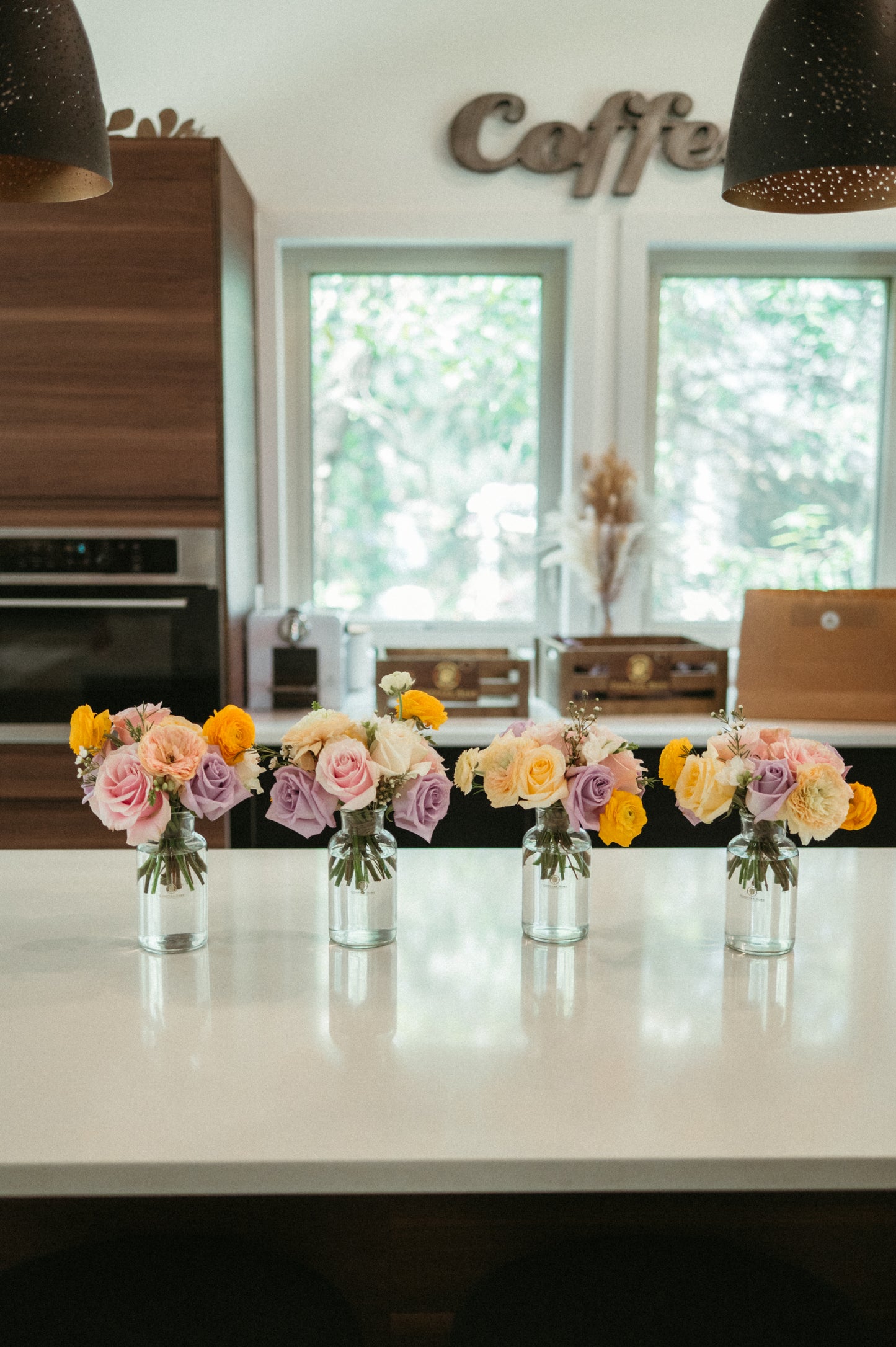 Decorative flowers in glass vases on a kitchen counter with 'Coffee' sign in the background.