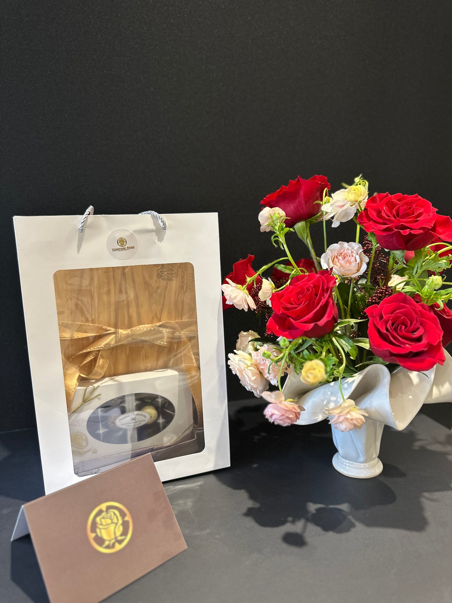Bouquet of red and white roses  in a handmade vase with a gift box, chocolates and card on a dark surface
