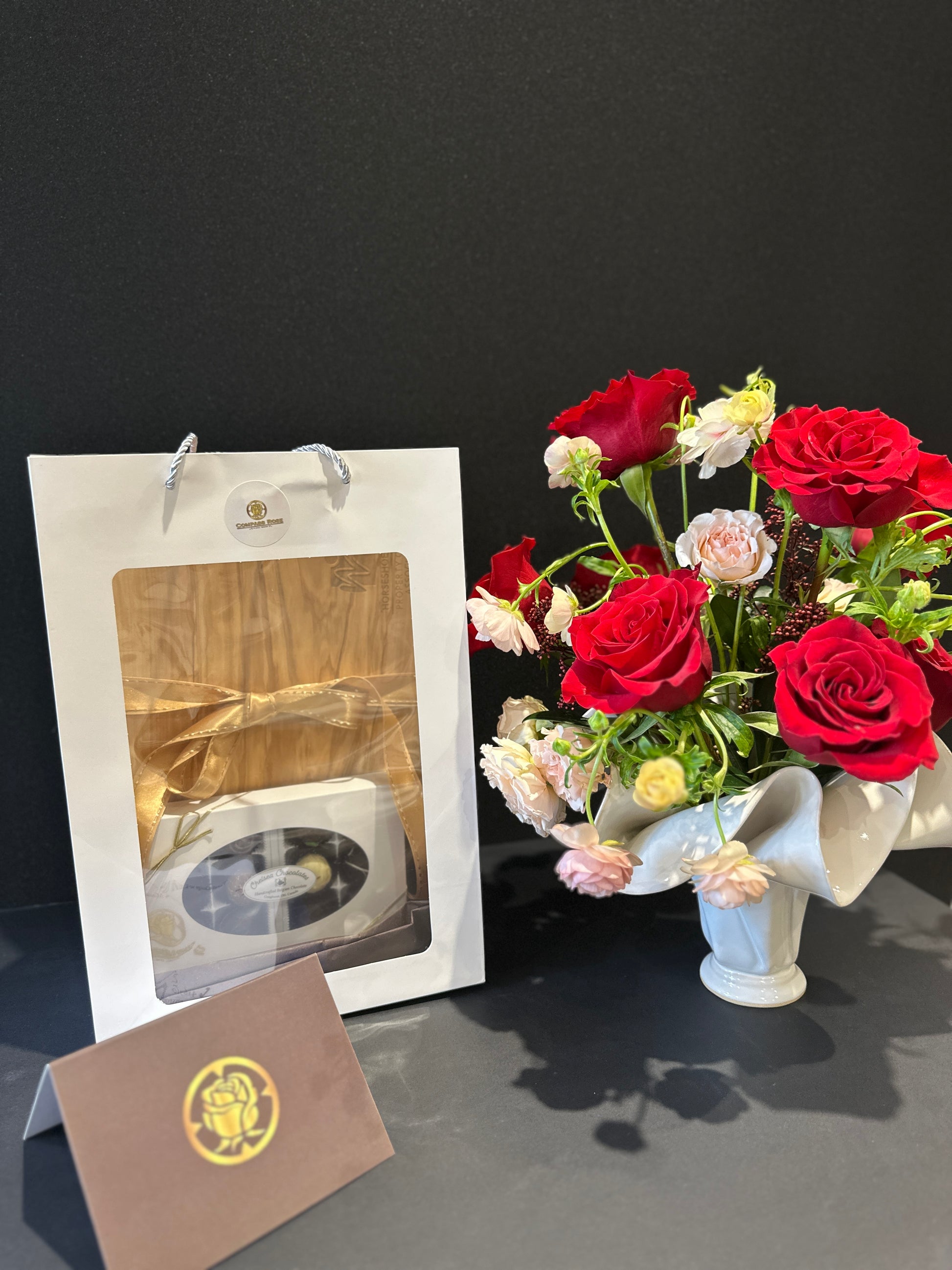 Bouquet of red and white roses  in a handmade vase with a gift box, chocolates and card on a dark surface