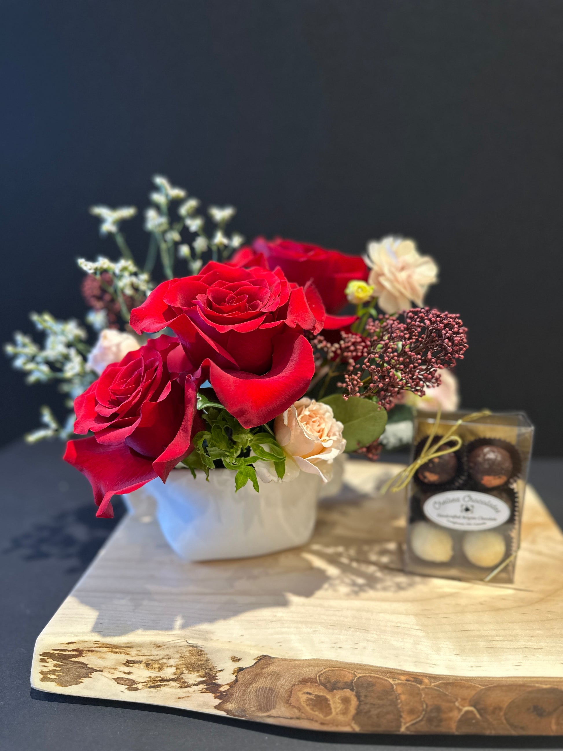 Bouquet of red roses and white flowers with a small box of chocolates on a charcuterie board.